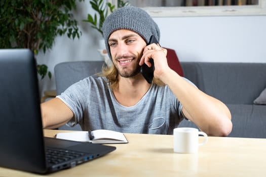 Handsome young caucasian man using computer working at home and talking with mobile feeling happy with smile showing teeth