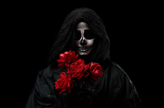 woman in a black hood with a skeleton make-up holds a bouquet of red roses in front of her, an invented image for the Halloween holiday