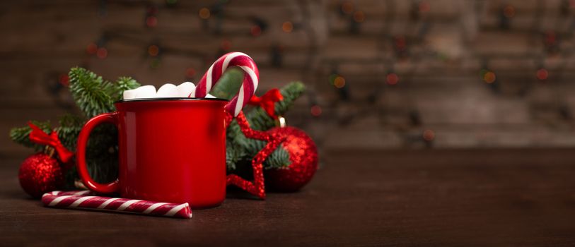 Cocoa hot chocolate in red mug with marshmallows candy cane fir tree branches and red baubles on dark wooden background