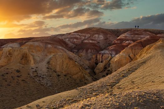 Beautiful low angle shot of two tiny human figures climbing massive red mountain in Kyzyl-Chin valley, also called Mars valley. Sunset cloudy sky as a backdrop. Golden hour. Altai, Siberia, Russia.