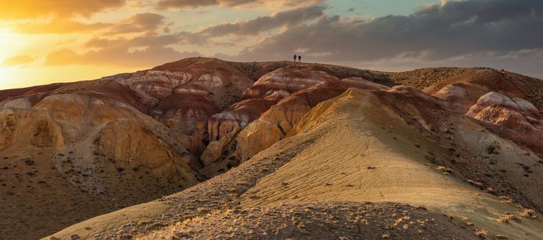 Low angle panoramic shot of two tiny figures on top of massive red mountain in Kyzyl-Chin valley, also called Mars valley. Beautiful sunset sky as a background. Golden hour. Altai, Siberia, Russia.