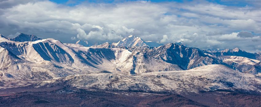 Scenic panoramic aerial view of snowy mountain peaks and slopes of North Chuyskiy ridge. Beautiful cloudy blue sky as a background. Altai mountains, Siberia, Russia.