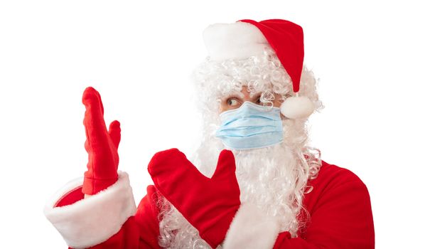 View of a man wearing a Santa Claus costume, medical mask and pointing with both hands at something next to him, isolated on white background. New normal, pandemic holiday concepts.
