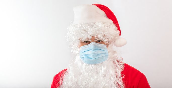View of a man wearing a Santa Claus costume and a medical mask on white background. Man looks stressed, angry, and agitated, staring straight in the camera. New normal, new reality, holiday concepts.