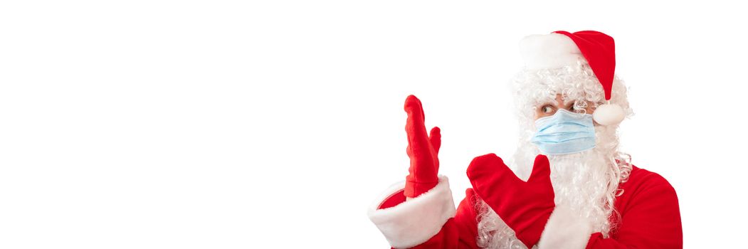 View of a man wearing a Santa Claus costume, medical mask and pointing with both hands at something next to him, isolated on white background. Banner size, copy space. Pandemic holiday concept.