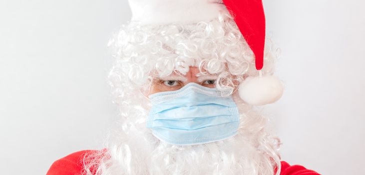 Close-up shot of man wearing a Santa Claus costume and a medical mask on white background. Man looks very angry and stressed, staring straight in the camera. New normal, new reality, holiday concepts