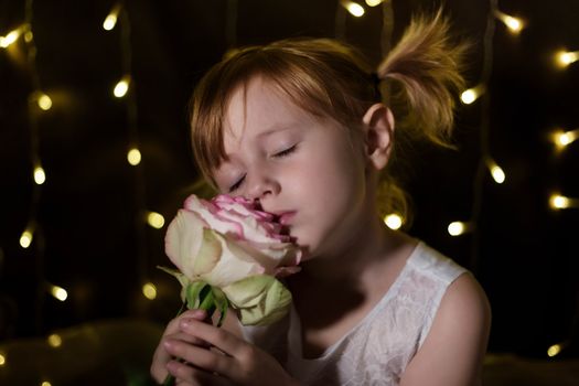 Adorable little girl with pink rose flower is posing in christmas lights on dark background