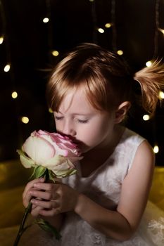 Cute lovely little girl with pink rose flower over dark background with garlands bokeh