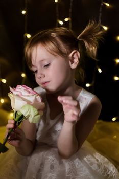 Adorable little girl with pink rose flower is posing in christmas lights on dark background