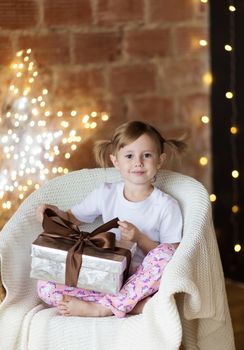 Beautiful child with a christmas gift in pajamas sitting on the chair with gift box looking at the camera