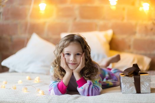 Adorable girl in pajamas lies on the bed and looks at the camera against the background of New Year's garlands