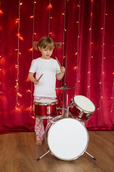 Girl in pajamas plays drums on red background with christmas garlands