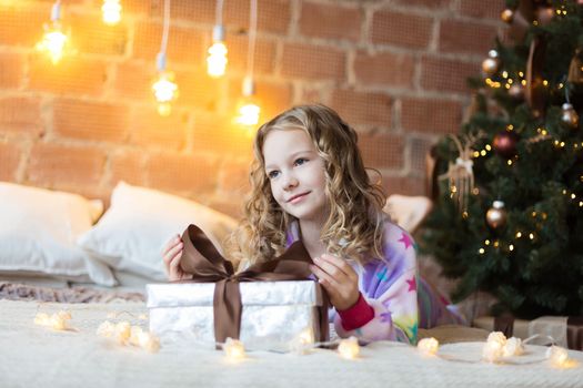 Cute Girl in pajamas lies on bed with a gift box and new year tree and lights behind