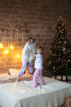 .Little sisters in pajamas on the bed fight with pillows against the background of christmas garlands