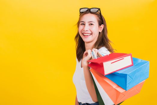 Asian Thai portrait happy beautiful cute young woman smiling stand with sunglasses excited holding shopping bags multi color looking camera, studio shot isolated yellow background with copy space
