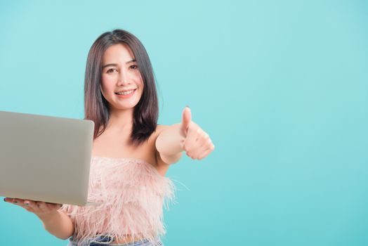 Asian happy portrait beautiful young woman standing smile her  holding laptop computer show finger thumb and looking to side on blue background with copy space for text