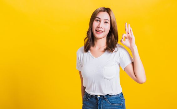 Asian Thai happy portrait beautiful cute young woman standing wear t-shirt showing gesturing ok sign with fingers looking to camera, isolated studio shot on yellow background with copy space