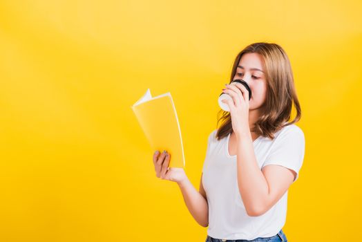 Portrait Asian Thai beautiful happy young woman standing holding drinking take away cup coffee paper and reading yellow book or diary, studio shot isolated on yellow background, with copy space