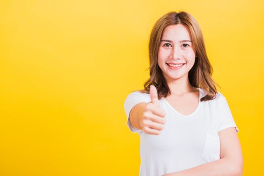 Portrait Asian Thai beautiful happy young woman smiling wear white t-shirt standing successful woman giving one thumbs up gesture sign in studio shot, isolated on yellow background with copy space