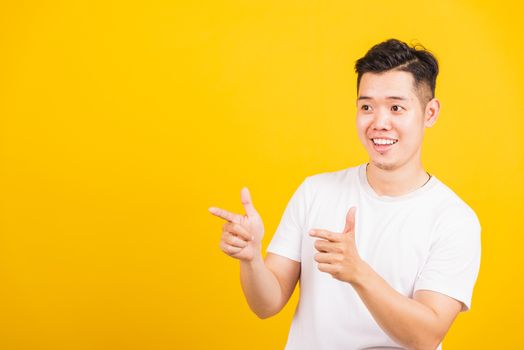 Portrait happy Asian handsome young man smiling standing wearing white t-shirt pointing finger to the side away he looking to space, studio shot isolated yellow background