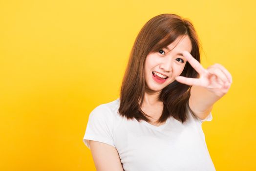 Asian happy portrait beautiful cute young woman teen smile standing wear t-shirt showing finger making v-sign symbol near eye  looking to camera isolated, studio shot yellow background with copy space