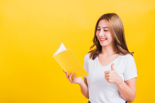 Portrait Asian Thai beautiful happy young lifestyle woman stands holding yellow book or diary she show thumb up finger and looking to book, studio shot isolated on yellow background, with copy space