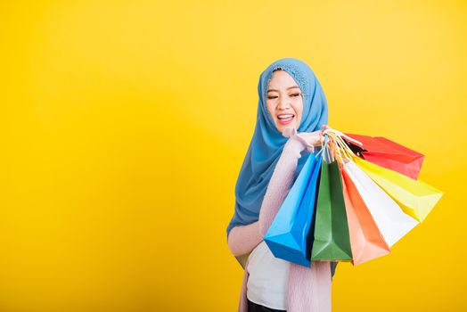 Asian Muslim Arab, Portrait of happy beautiful young woman Islam religious wear veil hijab funny smile she holding colorful shopping bags so glad shopping bags hand raise in studio isolated on yellow