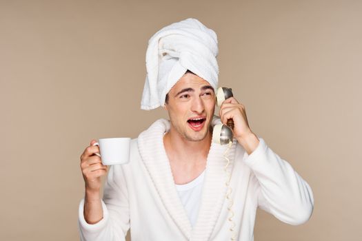 A man with a towel on his head is talking on the phone and holding a cup of coffee in his hand