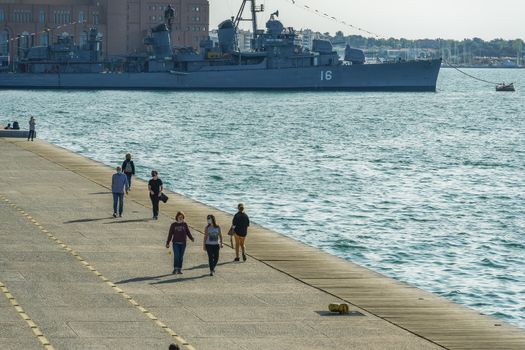 Unidentified persons with face protection walk on the pedestrian area of the city waterfront.