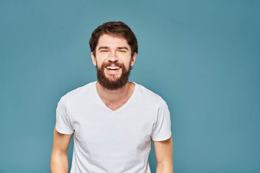 A bearded man in a white T-shirt gestures with his hands emotions blue background. High quality photo