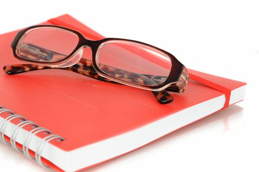 A closeup shot of a red notebook and pair of eye glasses over a white background.
