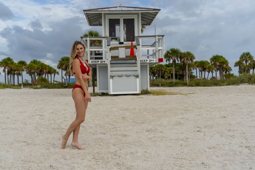 A beautiful blonde bikini model enjoys the weather outdoors on the beach while posing near a lifeguard station