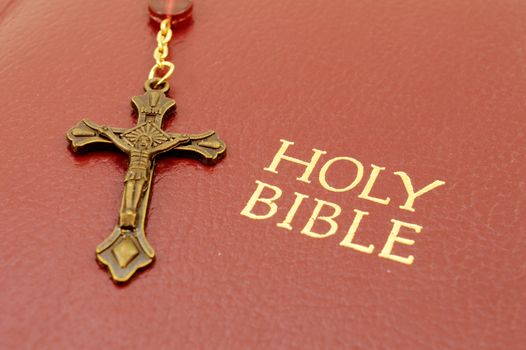 A closeup of a rosary cross and the Holy Bible book title printed in gold foil.