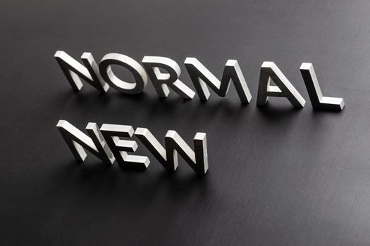 words new normal standing up with silver metal letters on flat black matte surface in tilted perspective with dark lightiing