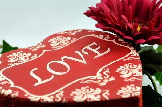 A closeup image of a red heart shaped box with the word love spelled on the lid and a maroon flower with greens for the romantic gift exchange.
