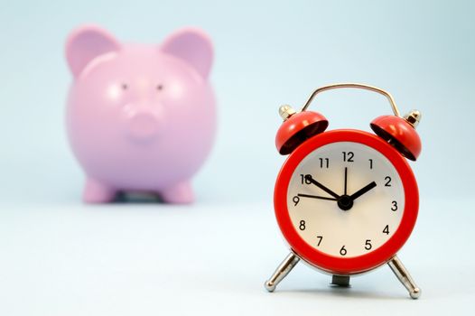 A red alarm clock focused in the forground with a blurred pink piggy coin bank in the background all over blue.