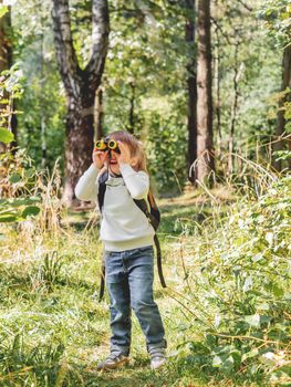 Curious boy is hiking in forest. Outdoor leisure activity for kids. Child looks through binoculars on tree foliage. Sunny day at autumn or summer day.