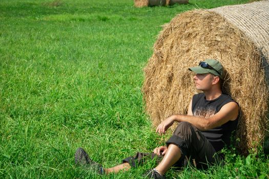 Sitting man to the green meadow, based on a hay bale, summer sunny day