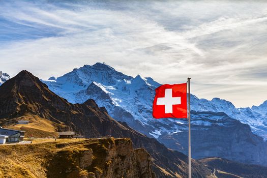 Stunning view of famous Jungfrau of Swiss Alps on Bernese Oberland from Mannlichen station, with Swiss national flag in foreground, on a sunny autumn day, Canton of Bern, Switzerland
