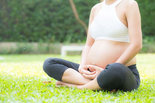 Beautiful pregnant asian young woman relax in the park, girl with belly sitting with crossed leg on grass and yoga exercise for wellness, copy space.