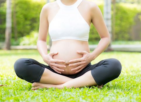 Beautiful pregnant asian young woman relax in the park, girl with belly sitting with crossed leg on grass and yoga exercise for wellness, copy space.