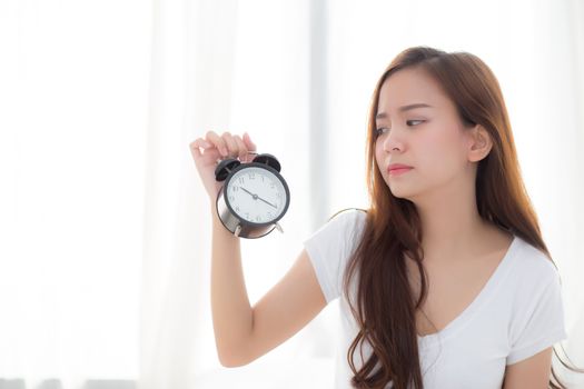 Beautiful young asian woman wake up in morning annoyed alarm clock holding hand, girl standing hurry wake late with appointment with curtain background on bedroom, lifestyle concept.