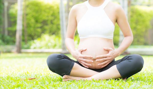 Beautiful pregnant asian young woman relax in the park, girl with belly sitting with crossed leg on grass and yoga exercise for wellness, copy space.