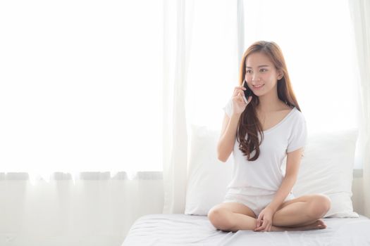 Beautiful of portrait young asian woman talking smart mobile phone and smile sitting at curtain background on bedroom, girl calling telephone, communication concept.