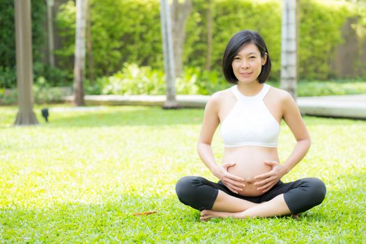Beautiful pregnant asian young woman relax in the park, girl with belly sitting with crossed leg on grass and yoga exercise for wellness, copy space.