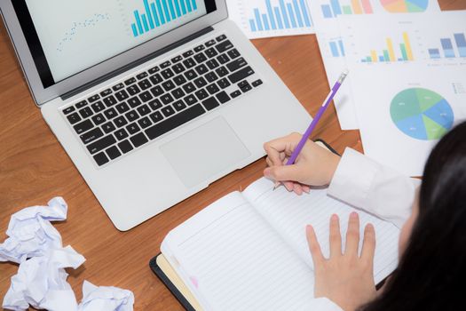 Businesswoman writing on notebook at desk, asian woman with not idea with graph analysis laptop and crumpled paper at office, freelance business concept.
