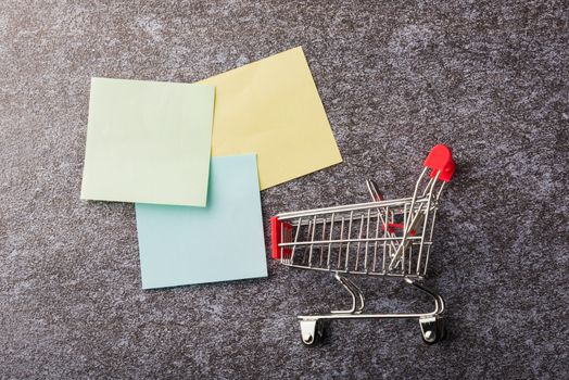 Above blank paper stick note list yellow and green with the shopping cart on concrete background, Shopping reminder concept