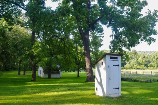 Beautiful white colonial outhouse in natural light by a stone cottage. Idllic village setting with tall trees, green grass, fences and copy space.