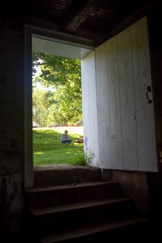 Full frame image of female college student reading / studying outside, sitting in the grass on a sunny summer day, seen trough a rusric wooden door.