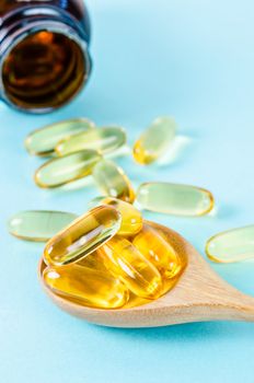 Fish oil capsules in wooden spoon with bottle on blue background.